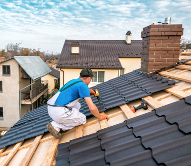 In the image, there is a person working on a roof construction project, wearing a blue shirt and white pants, with tools and materials around them. The roof is covered with black shingles, and there are houses in the background.