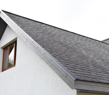 The image shows a residential building with a sloped roof covered in dark shingles, featuring a visible gutter system.
