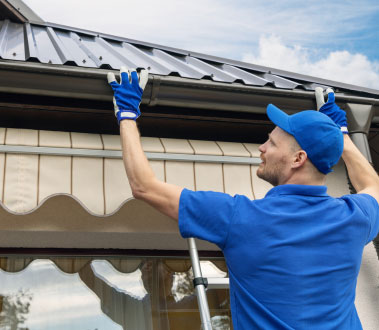 A man wearing blue gloves is working on a roof repair while standing on an extension ladder.