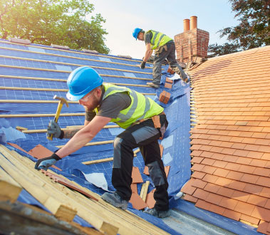 The image shows two construction workers engaged in roofing work on a partially constructed house, with one worker using a hammer and the other handling materials, under clear skies.