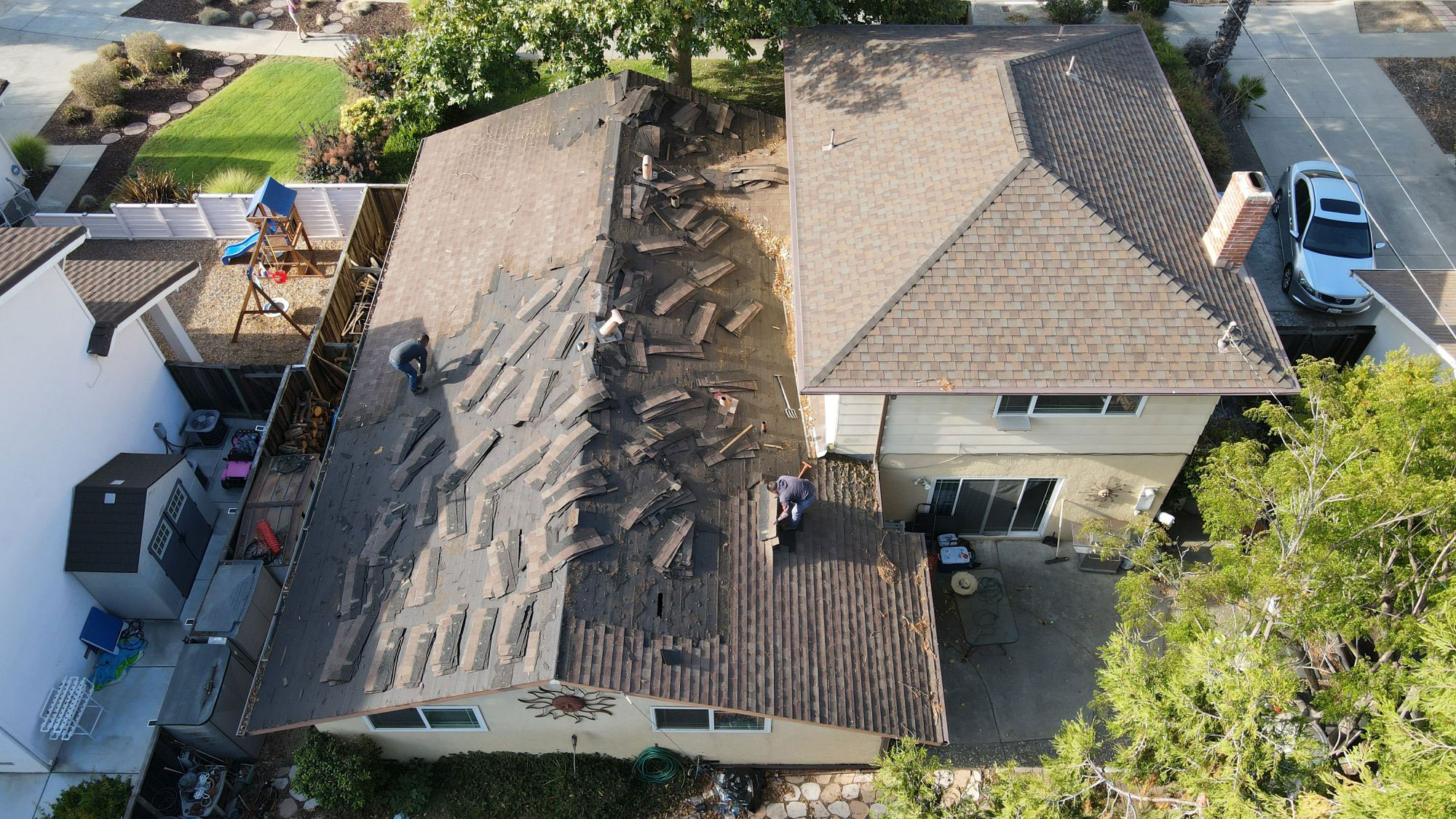 The image shows a residential house with a roof under construction or repair, featuring visible materials like shingles and plywood, and there are workers on the roof.