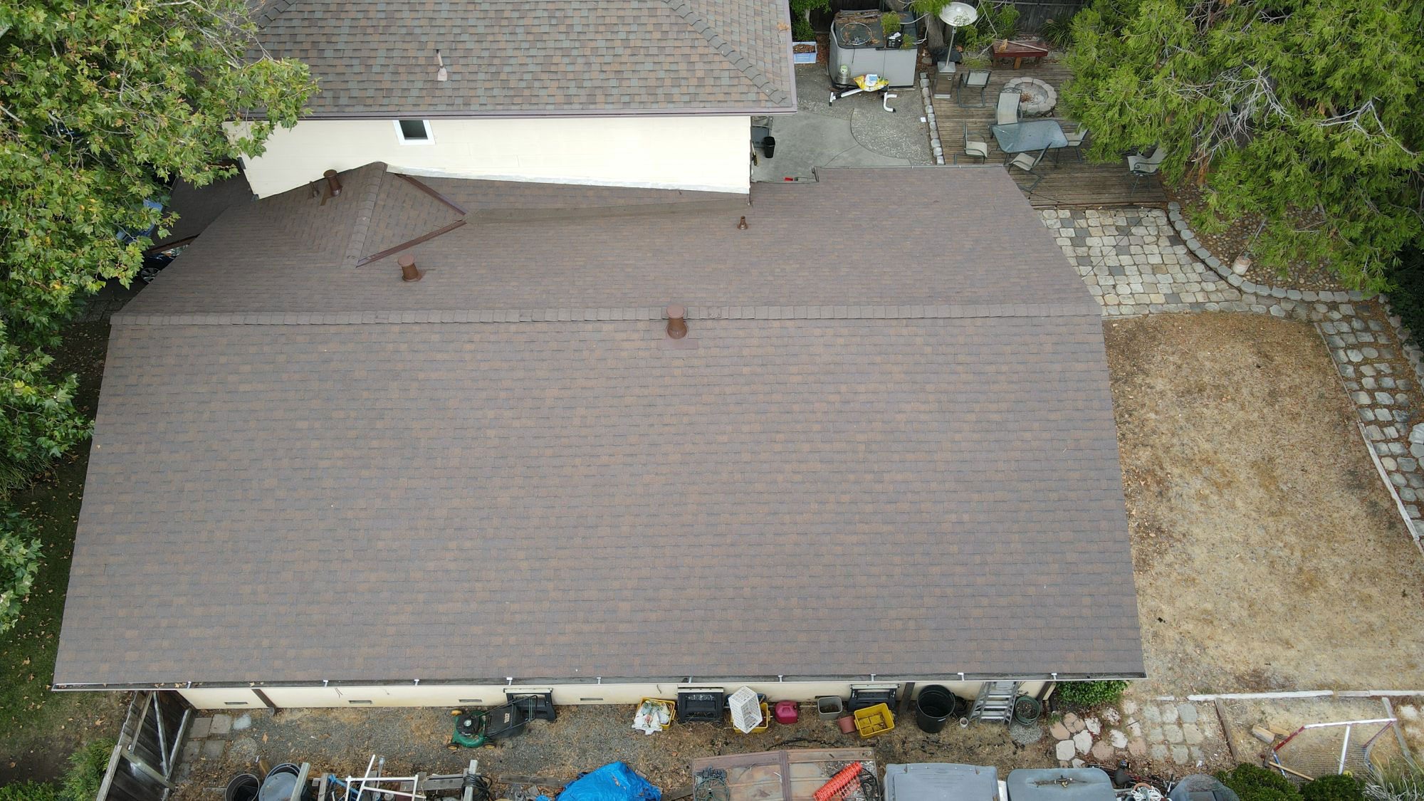 The image shows a residential house with a brown roof, surrounded by trees and a clear blue sky.