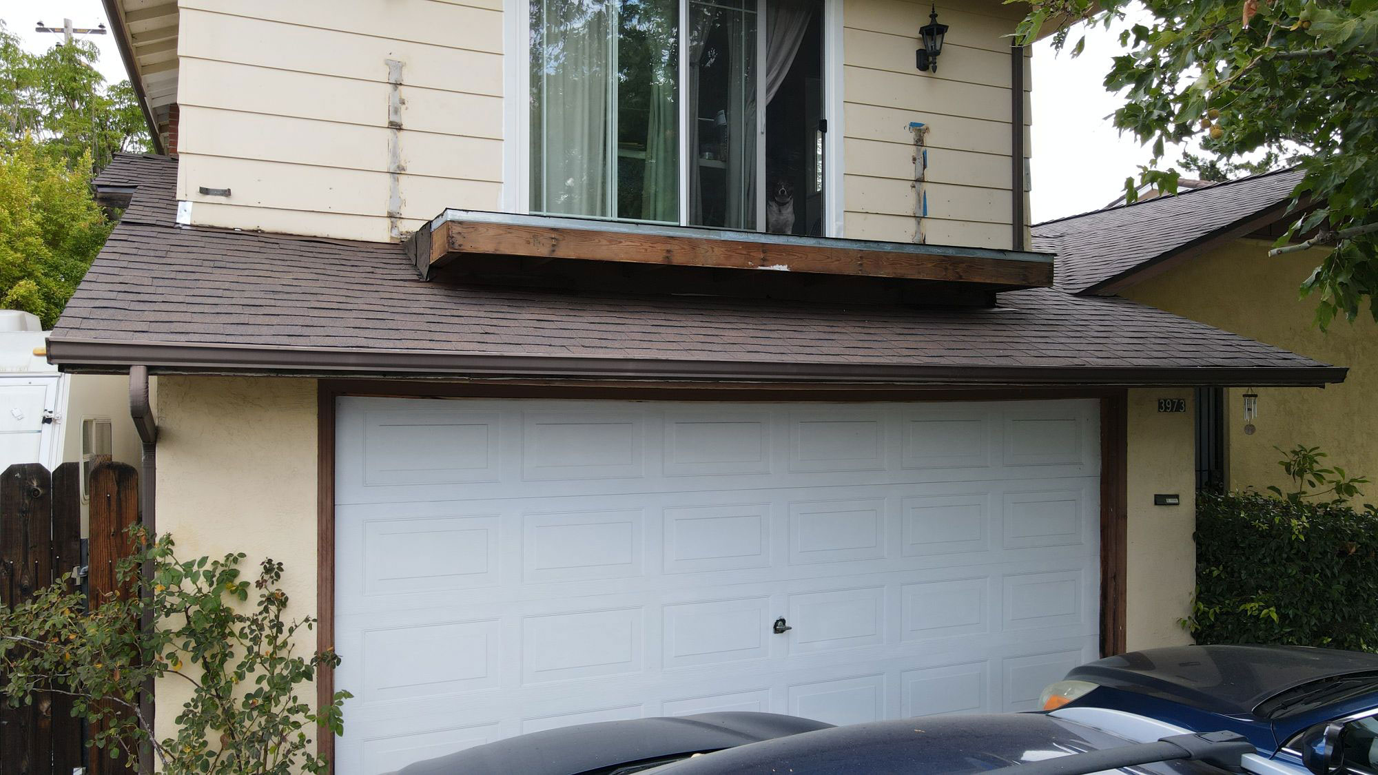 The image shows a two-story residential house with a garage door partially open, featuring a dark roof and a car parked on the driveway.