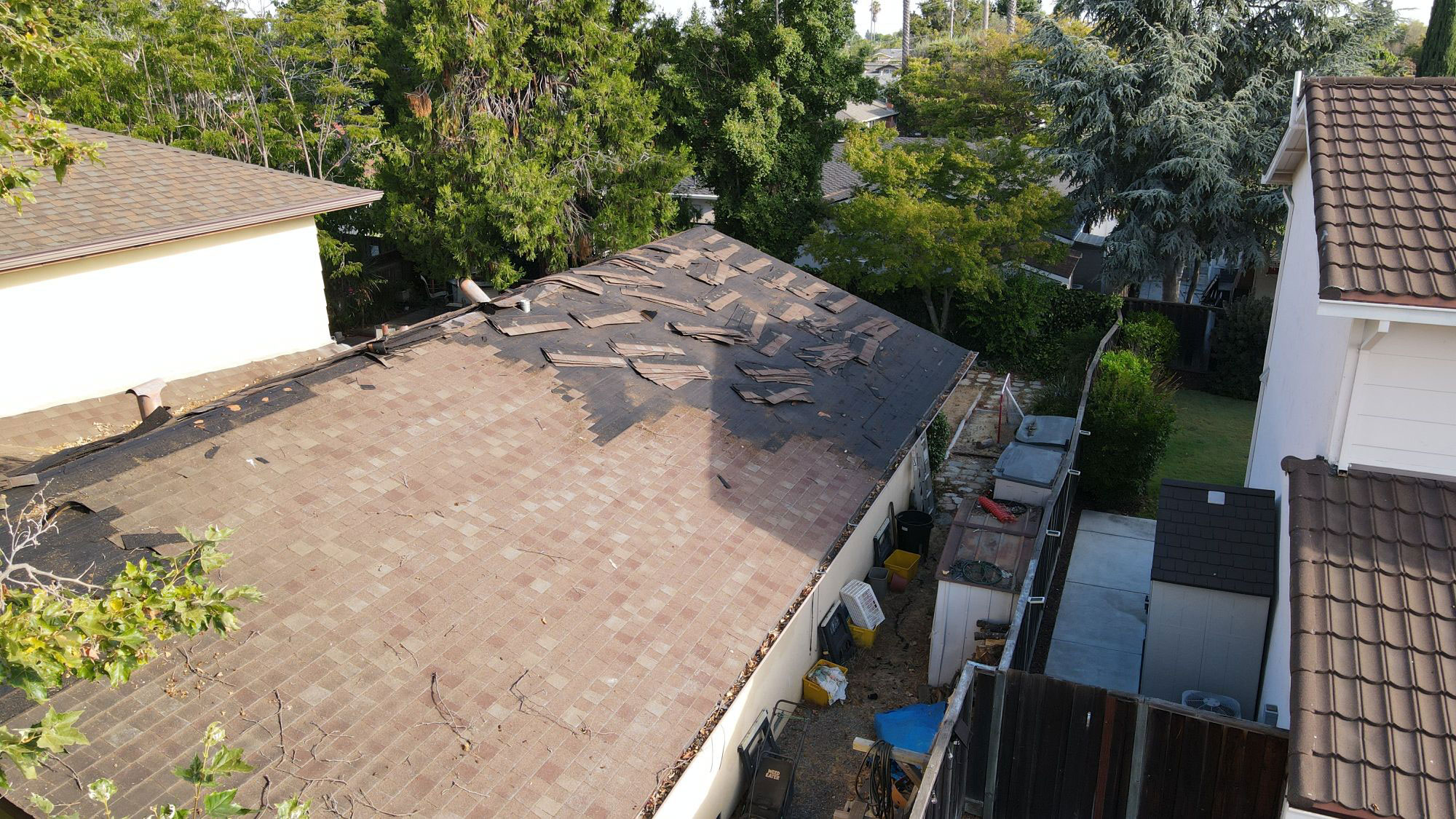 The image shows a residential house with a damaged roof undergoing repairs, viewed from an elevated angle, with a clear sky above and trees visible in the background.