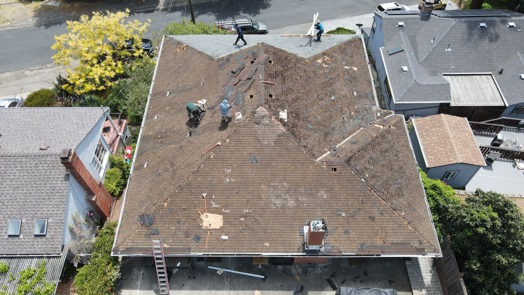 The image shows a residential building with a damaged roof under repair, featuring exposed shingles and construction materials, set against a suburban neighborhood backdrop.
