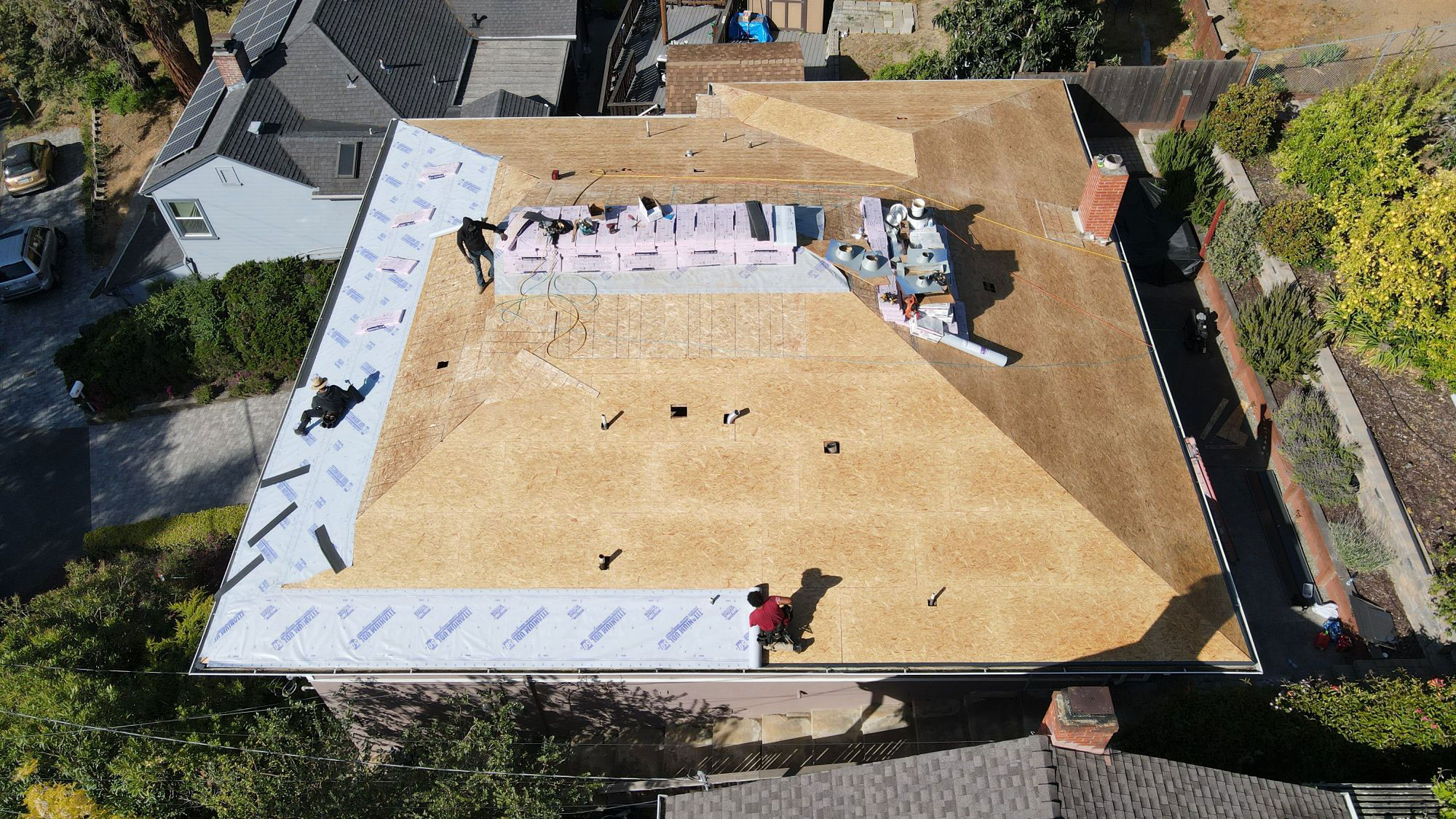 The image shows a house under construction with a visible foundation and framing, viewed from an angle that includes part of its roof and the surrounding environment, including a street and a building across the road.