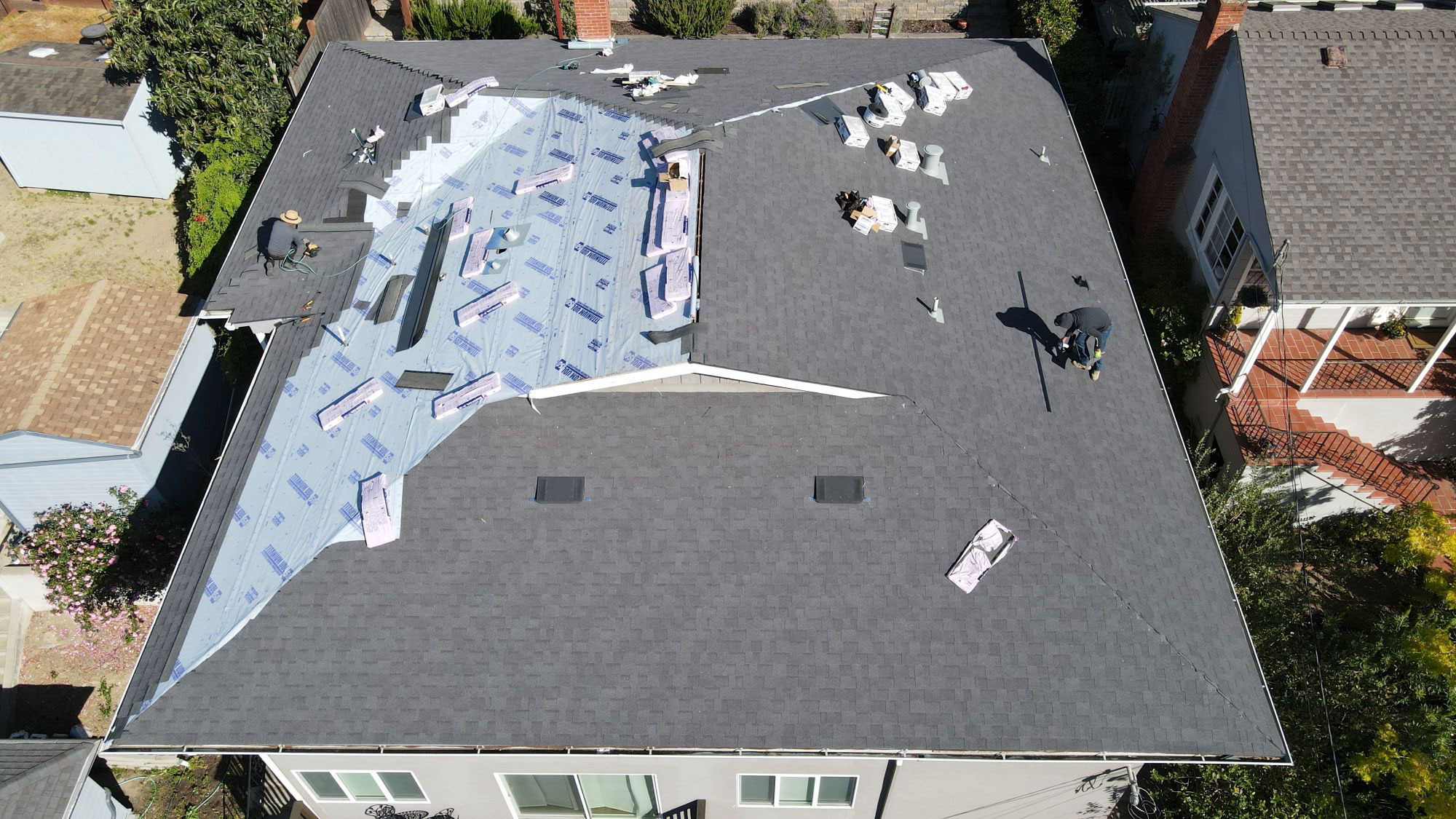 The image shows a residential roof undergoing repair or replacement, with visible tarps and construction materials on a sunny day.