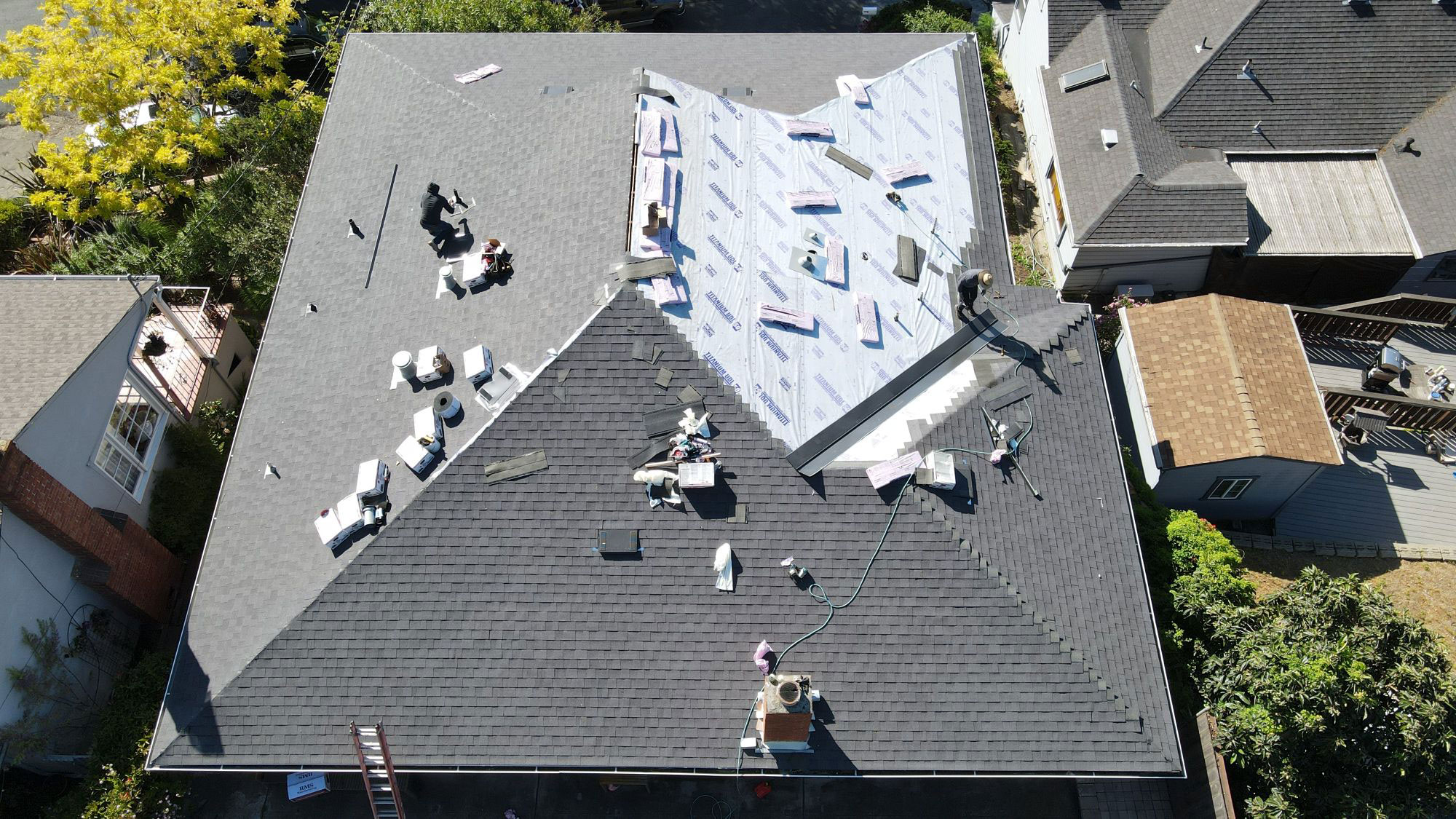 The image depicts a building under construction with a visible roofing material, surrounded by other buildings in a residential area.