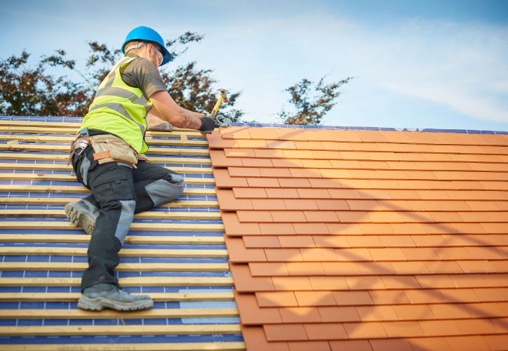 The image shows a construction worker on a roof, working on a tiled structure, with safety gear including a hard hat and harness.