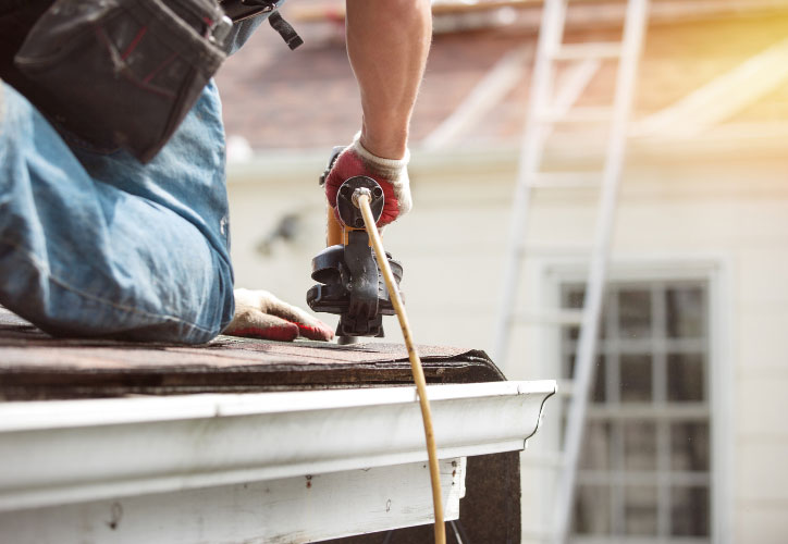 A construction worker using a drill on a wooden deck, with a focus on his hands and the drill, under a clear blue sky.