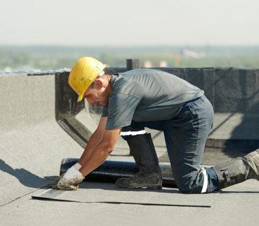 The image shows a construction worker crouched on a rooftop, working on installing roofing material with tools spread out around him.