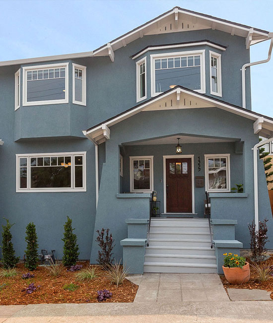 The image shows a two-story residential house with a prominent front porch, a garage door on the left side, and a well-maintained exterior with a mix of blue siding and white trim.