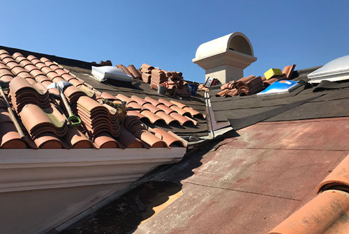 The image shows a rooftop with damaged tiles, some of which are missing, revealing the underlayment. The roof appears to be in need of repair.